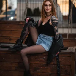 Kobieta z tatuażami siedzi na drewnianej ławce w czarnym topie, dżinsowych szortach i czarnej kurtce, pozując do zdjęcia z nogami w czarnych rajstopach.
