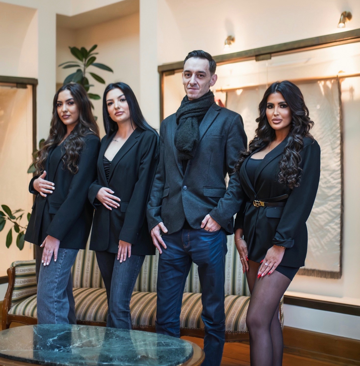 Grupa czterech osób pozuje w eleganckim wnętrzu, trzy kobiety w czarnych marynarkach i mężczyzna w szarym garniturze i szaliku, za nimi kanapa w paski i dekoracyjna ściana.