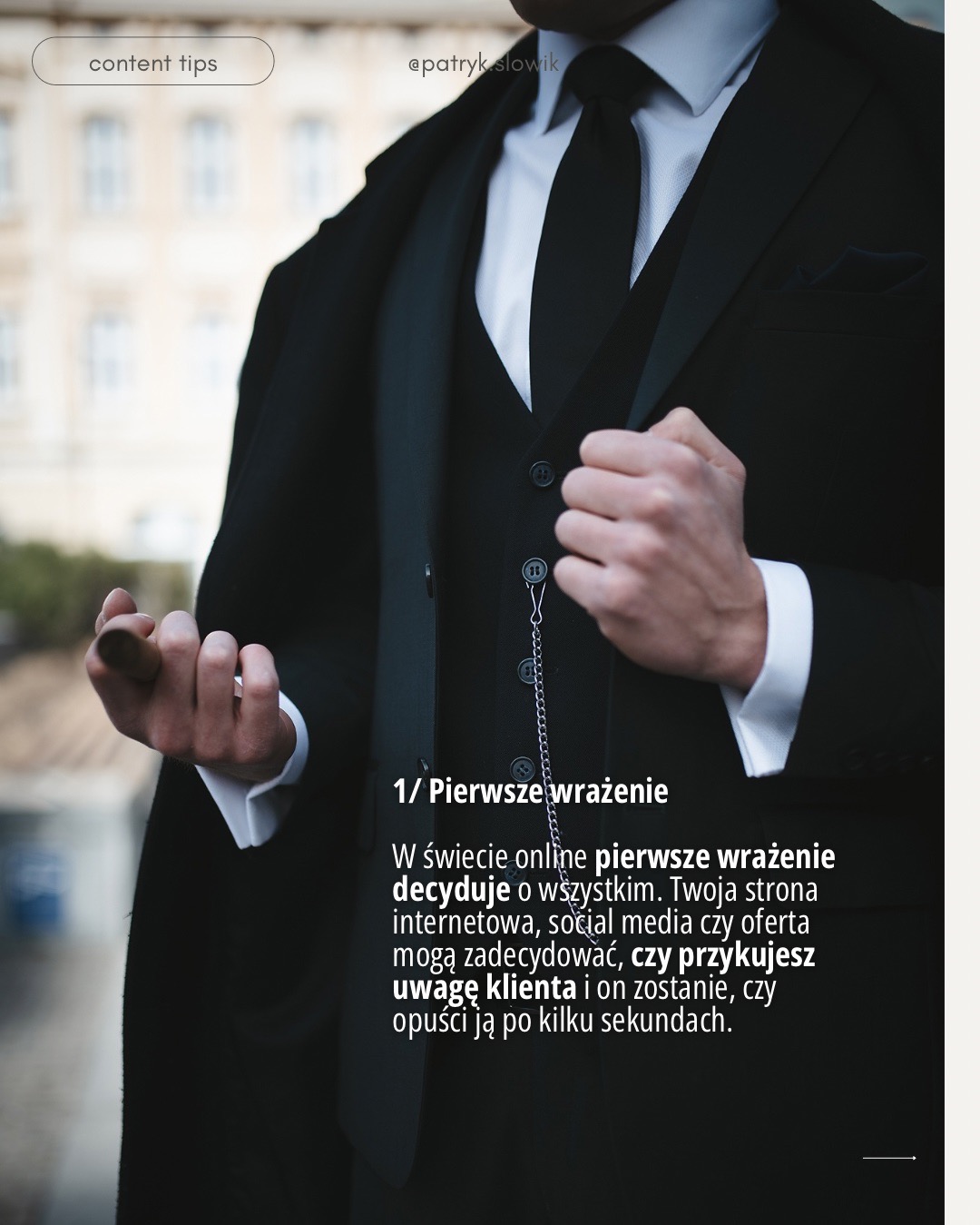 Mężczyzna w eleganckim, czarnym garniturze trzyczęściowym z łańcuszkiem od zegarka, prezentujący dłoń z palcami lekko zgiętymi, na rozmytym tle miejskiej architektury.