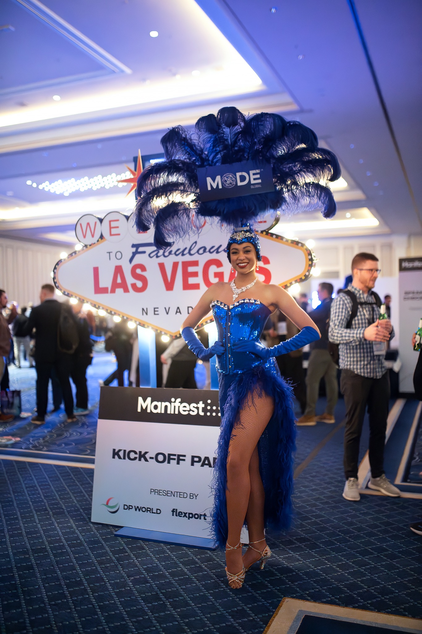 Kobieta w błyszczącym, niebieskim kostiumie z piórami na głowie pozuje przed znakiem 'Welcome to Fabulous Las Vegas' podczas eventu Manifest, uśmiechając się do kamery.