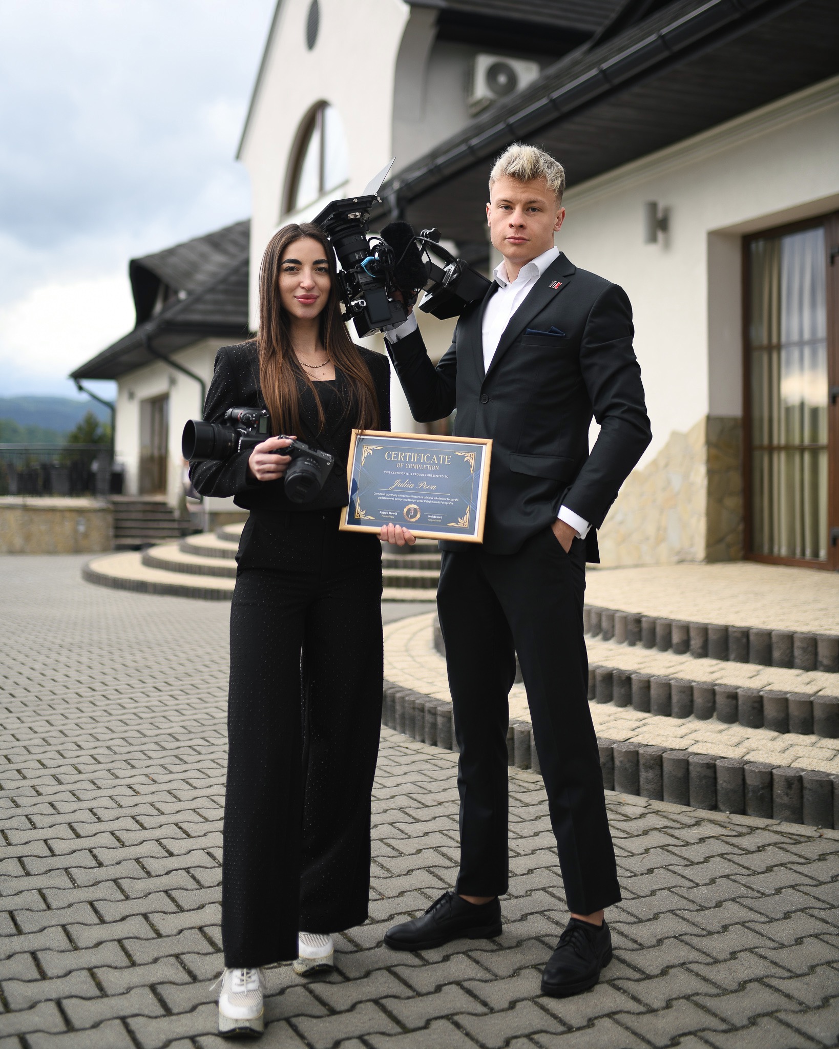 Dwoje elegancko ubranych fotografów z kamerami i certyfikatem na tle budynku z jasną elewacją i schodami. Kobieta trzyma aparat, mężczyzna kamerę.