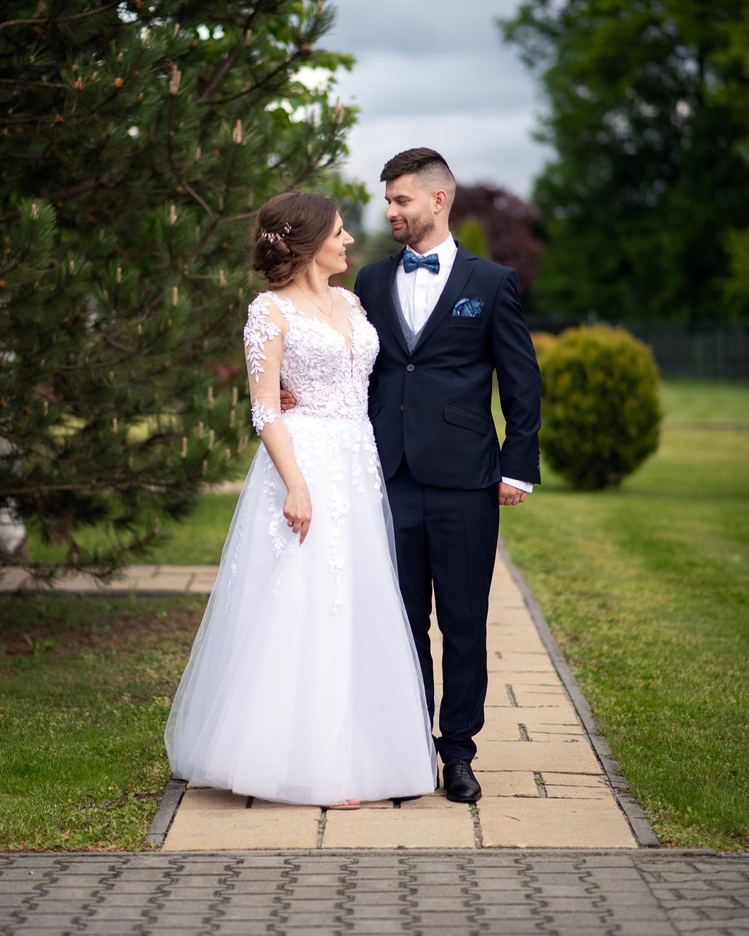 Uśmiechnięta para młoda w eleganckich strojach pozuje na tle zieleni. Panna młoda w sukni z koronkowymi rękawami, pan młody w granatowym garniturze z muchą.