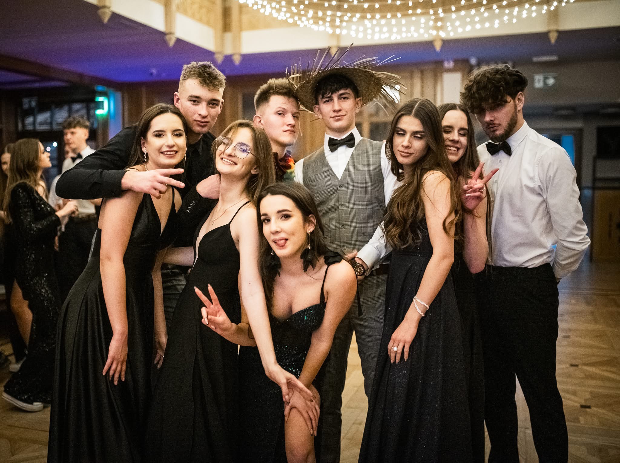 Grupa elegancko ubranych młodych ludzi pozuje do zdjęcia na tle sali balowej. Wśród nich osoba w słomkowym kapeluszu i muszce. Parkiet i dekoracje świetlne w tle.