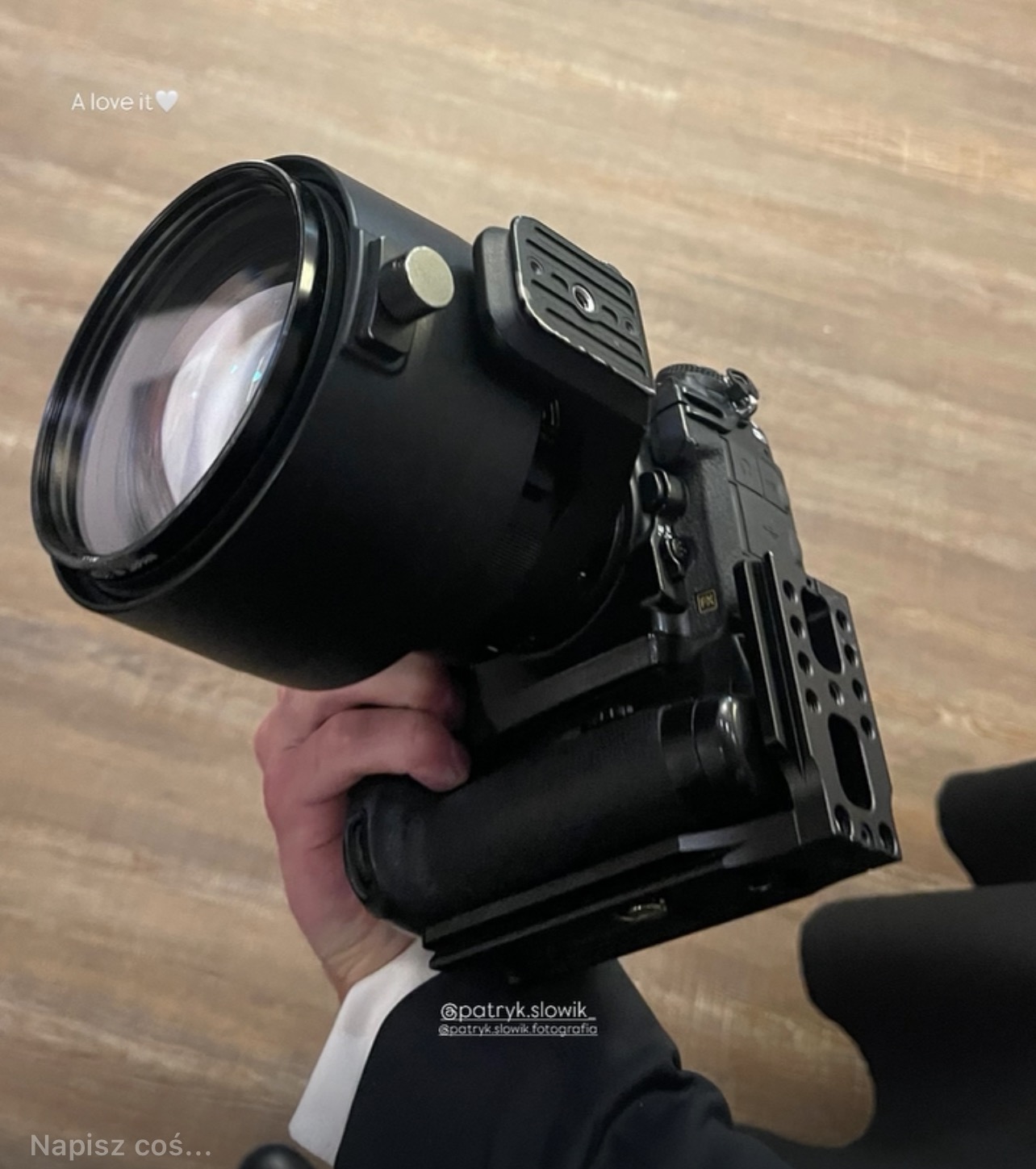 Czarny aparat z dużym obiektywem trzymany w dłoni, widoczny fragment białej koszuli i czarnego garnituru. Ujęcie z bliska, na jasnym tle.