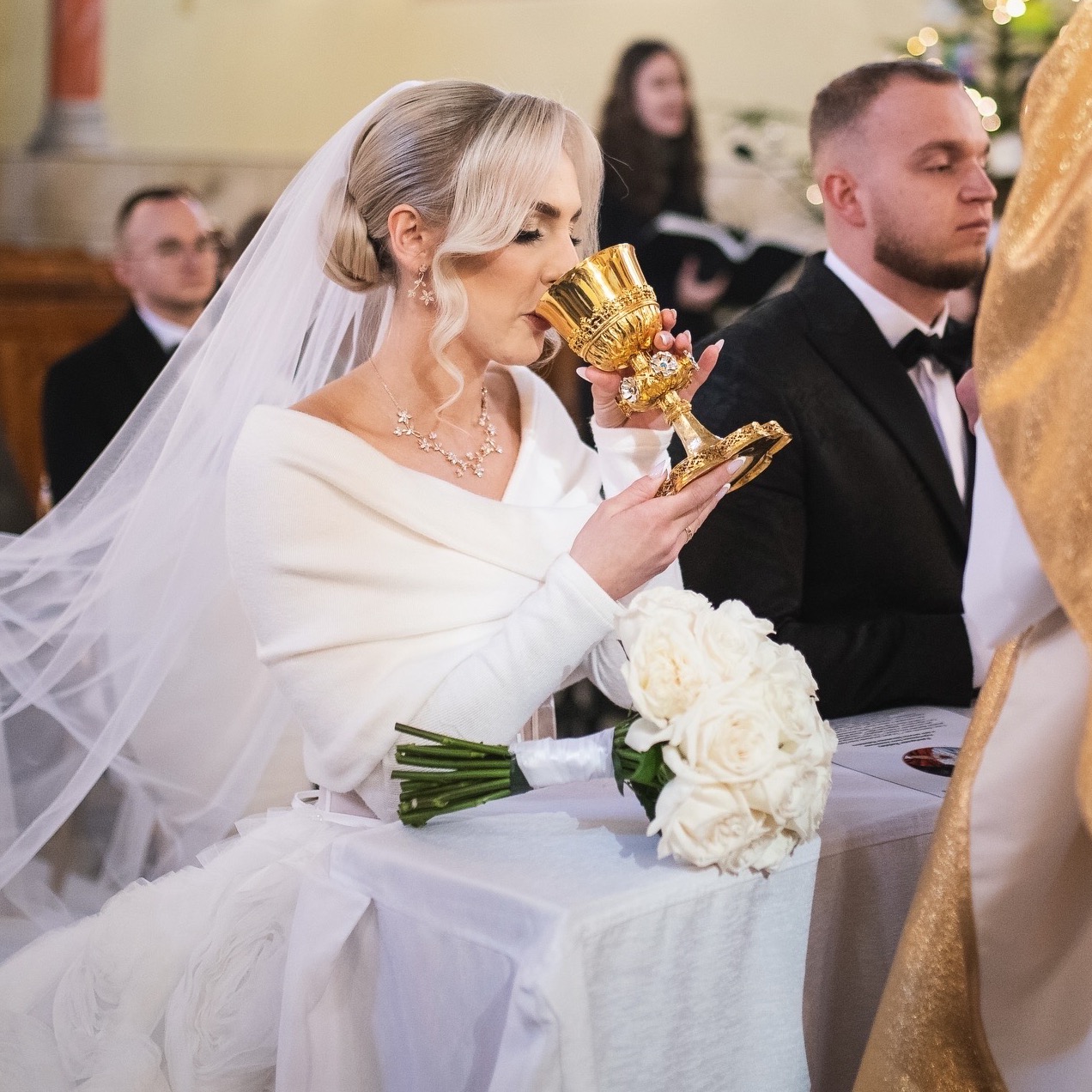 Panna młoda w białej sukni pije z bogato zdobionego, złotego kielicha podczas ceremonii ślubnej w kościele. W tle widoczny pan młody i inni goście.
