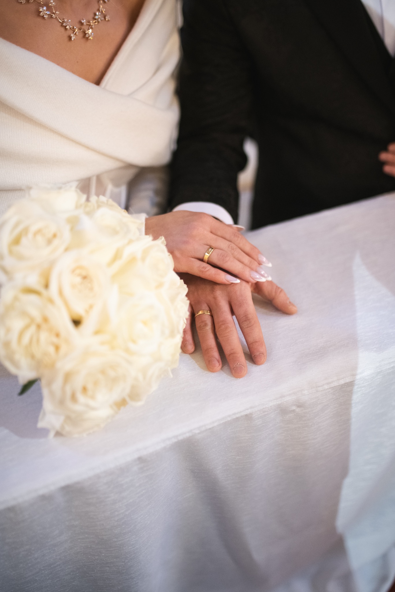 Dłonie pary młodej z obrączkami na białym obrusie obok bukietu białych róż. Kobieta w białym swetrze i naszyjniku, mężczyzna w czarnym garniturze.