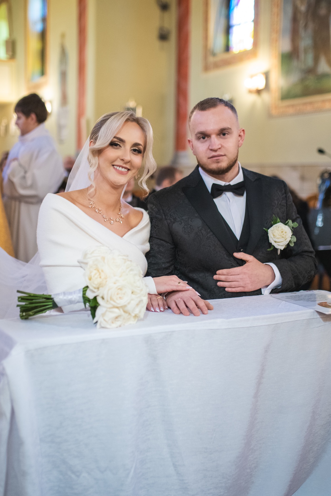 Uśmiechnięta para młoda w kościele, siedząca za stołem z bukietem białych róż. Panna młoda w białej sukni i welonie, pan młody w ciemnym garniturze z muszką.