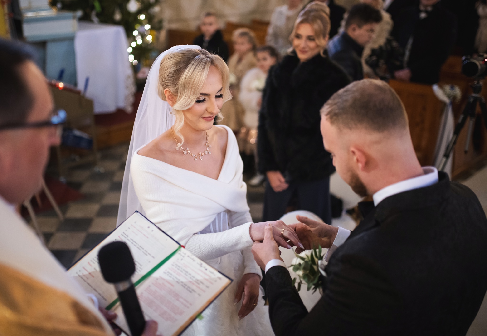 Moment zakładania obrączki podczas ceremonii ślubnej w kościele. Panna młoda w białej sukni, pan młody w czarnym garniturze. W tle goście i ksiądz z mikrofonem.