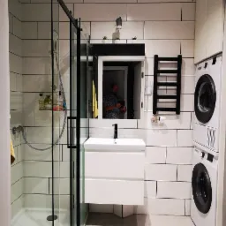 Nowoczesna łazienka z kabiną prysznicową typu walk-in z czarnymi ramami, białą umywalką nablatową z czarną armaturą, grzejnikiem drabinkowym i pralko-suszarką w zabudowie, wykończona białymi...