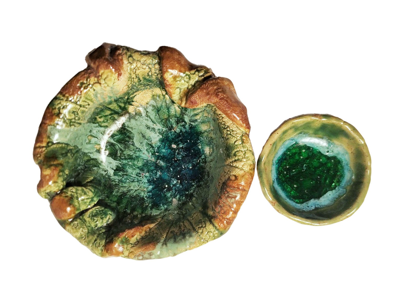 Dwie ceramiczne miseczki o nieregularnych kształtach i szkliwionej powierzchni, większa w odcieniach zieleni, brązu i błękitu, mniejsza zielona z jasnoniebieską obwódką, na białym tle.