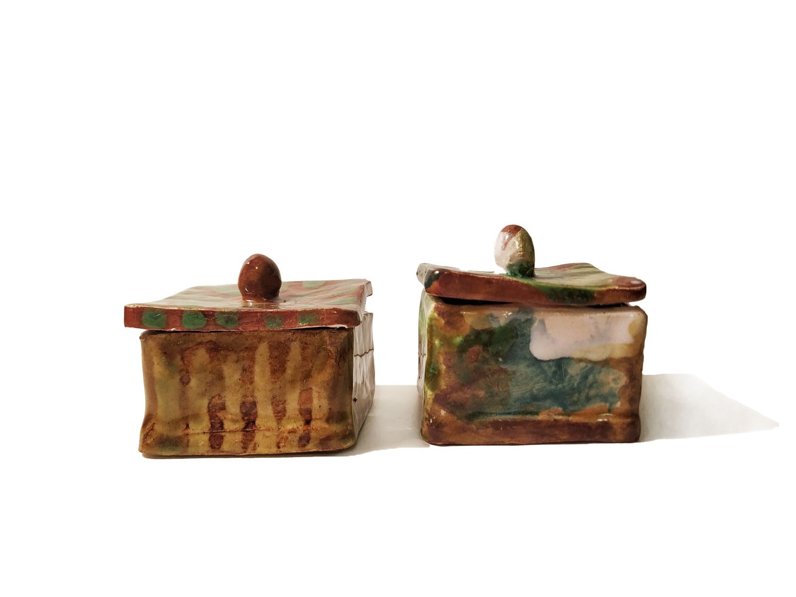 Dwa kwadratowe, ręcznie robione ceramiczne pojemniki z pokrywkami w odcieniach brązu, zieleni i bieli, każdy z unikalnym wzorem i fakturą, ustawione obok siebie na białym tle.