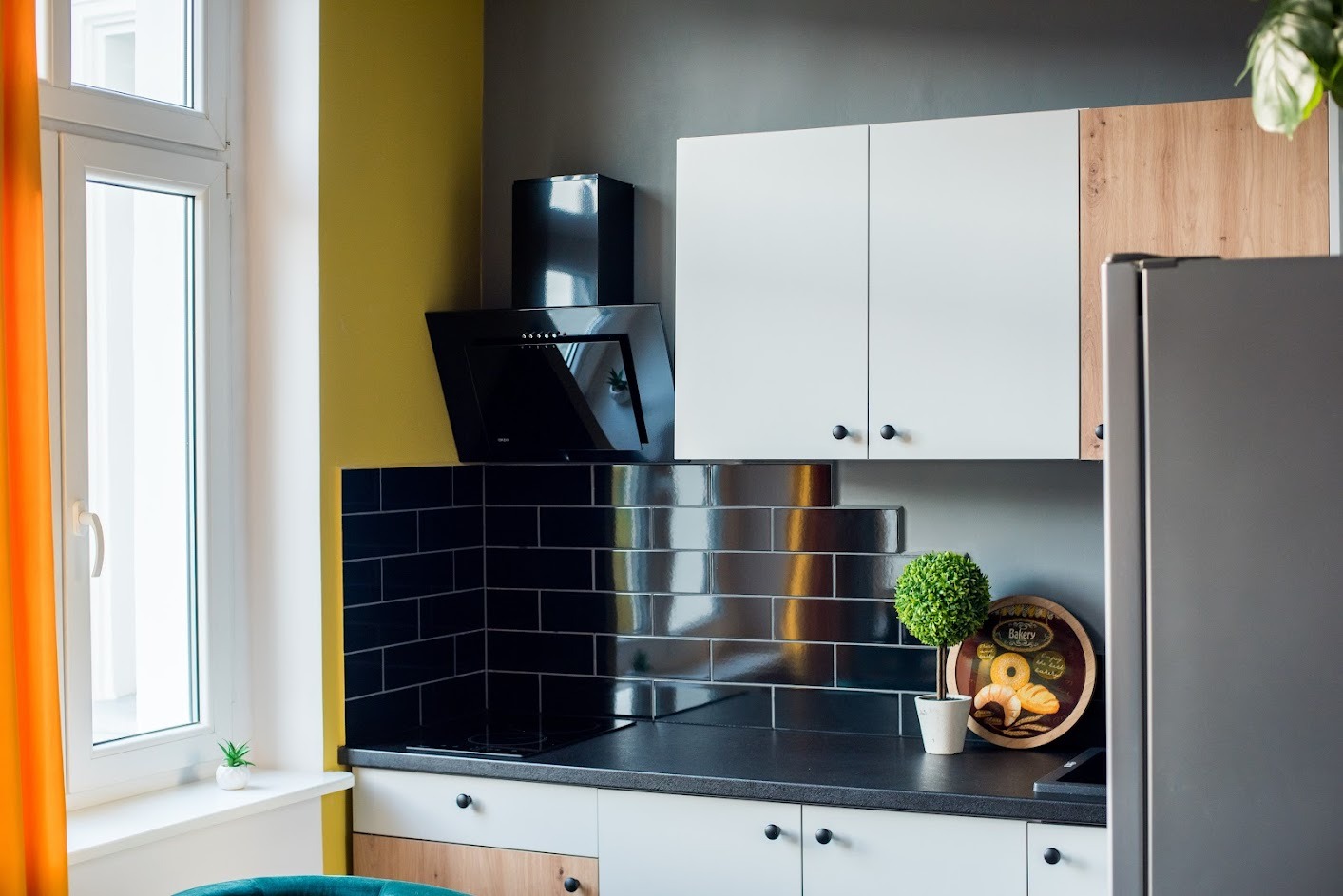 Nowoczesna kuchnia z czarnymi błyszczącymi płytkami ułożonymi w cegiełkę, białymi szafkami z okrągłymi czarnymi uchwytami, czarnym okapem i blatem, z akcentami w kolorze żółtym i drewnopodobnym...