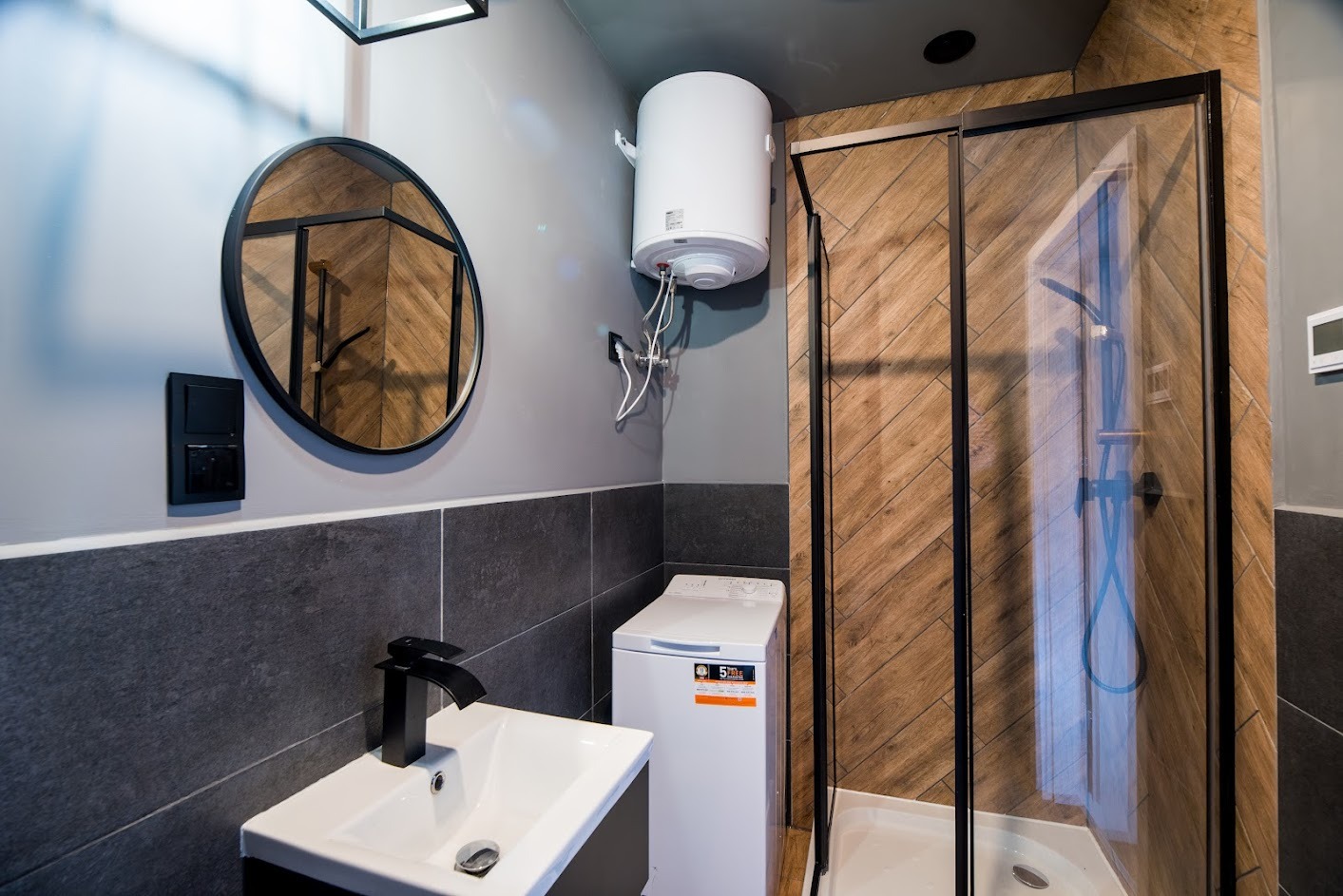 Nowoczesna łazienka z kabiną prysznicową z czarnymi ramami, okrągłym lustrem z czarną ramą, białą umywalką z czarną armaturą, pralką i bojlerem elektrycznym na szarej ścianie z ciemnymi płytkami.