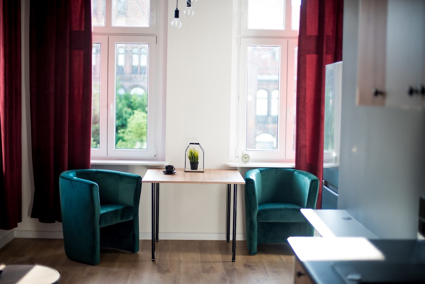 Jasne wnętrze z dwoma oknami z bordowymi zasłonami, pomiędzy którymi stoi mały stolik z czarnymi nogami, na nim roślina w czarnej doniczce i filiżanka, a po bokach dwa turkusowe fotele.