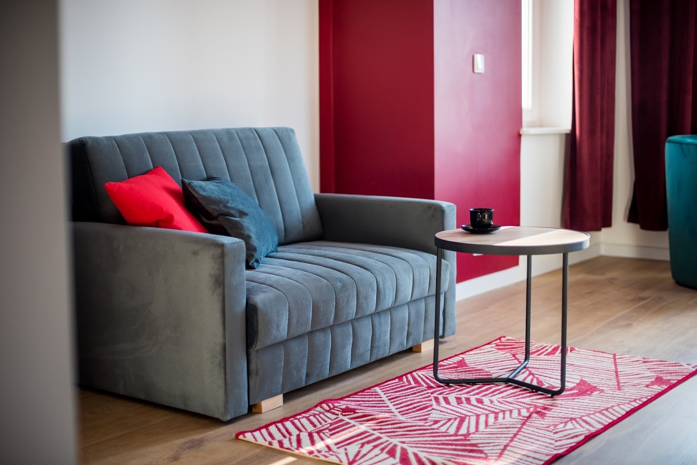 Szary, welurowy fotel z czerwoną i granatową poduszką stoi obok okrągłego stolika z czarną filiżanką, na drewnianej podłodze leży czerwono-biały dywanik, ściana w kolorze burgundu.