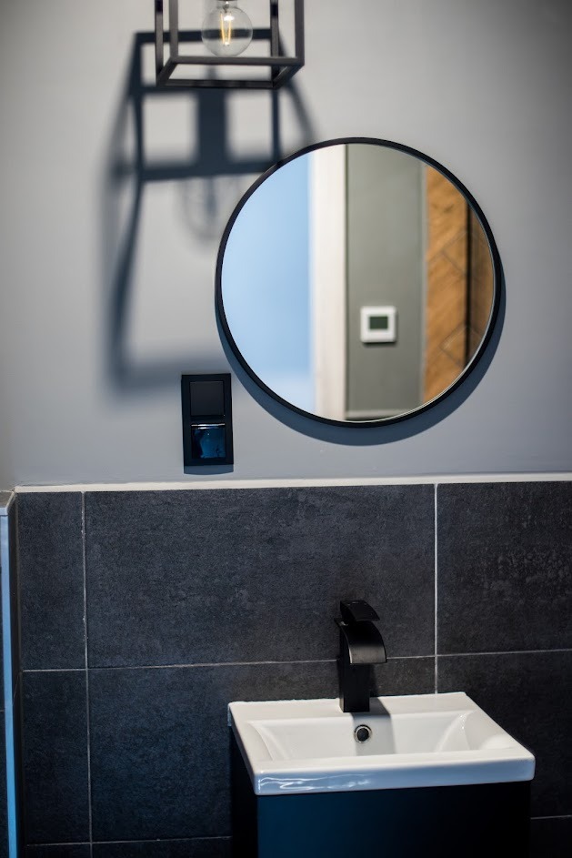 Nowoczesna łazienka z okrągłym lustrem, czarną armaturą i szarą glazurą, oświetlona lampą z żarówką edisona.