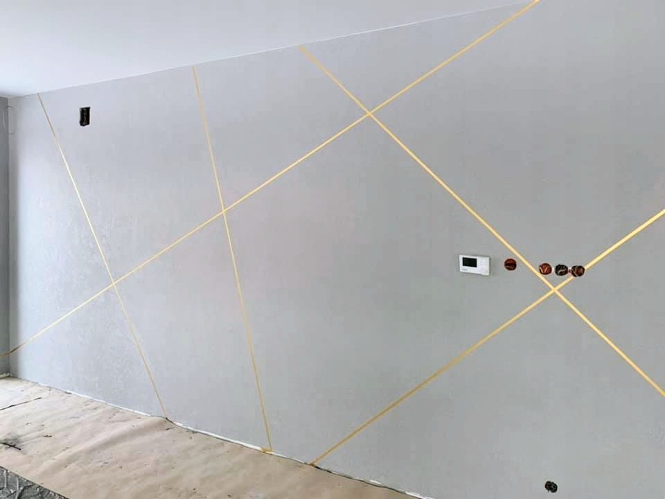 Szara ściana z dekoracyjnymi, złotymi, ukośnymi paskami, widoczne gniazdka elektryczne i niezabezpieczona podłoga.