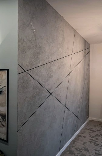 Ściana w salonie pokryta szarym tynkiem dekoracyjnym z czarnymi, geometrycznymi liniami, widoczne wystające kable elektryczne w górnym lewym rogu.