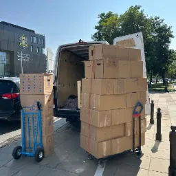 Załadunek kartonowych pudeł na wózkach do białego busa na tle budynku Warszawskiego Towarzystwa Wioślarskiego.