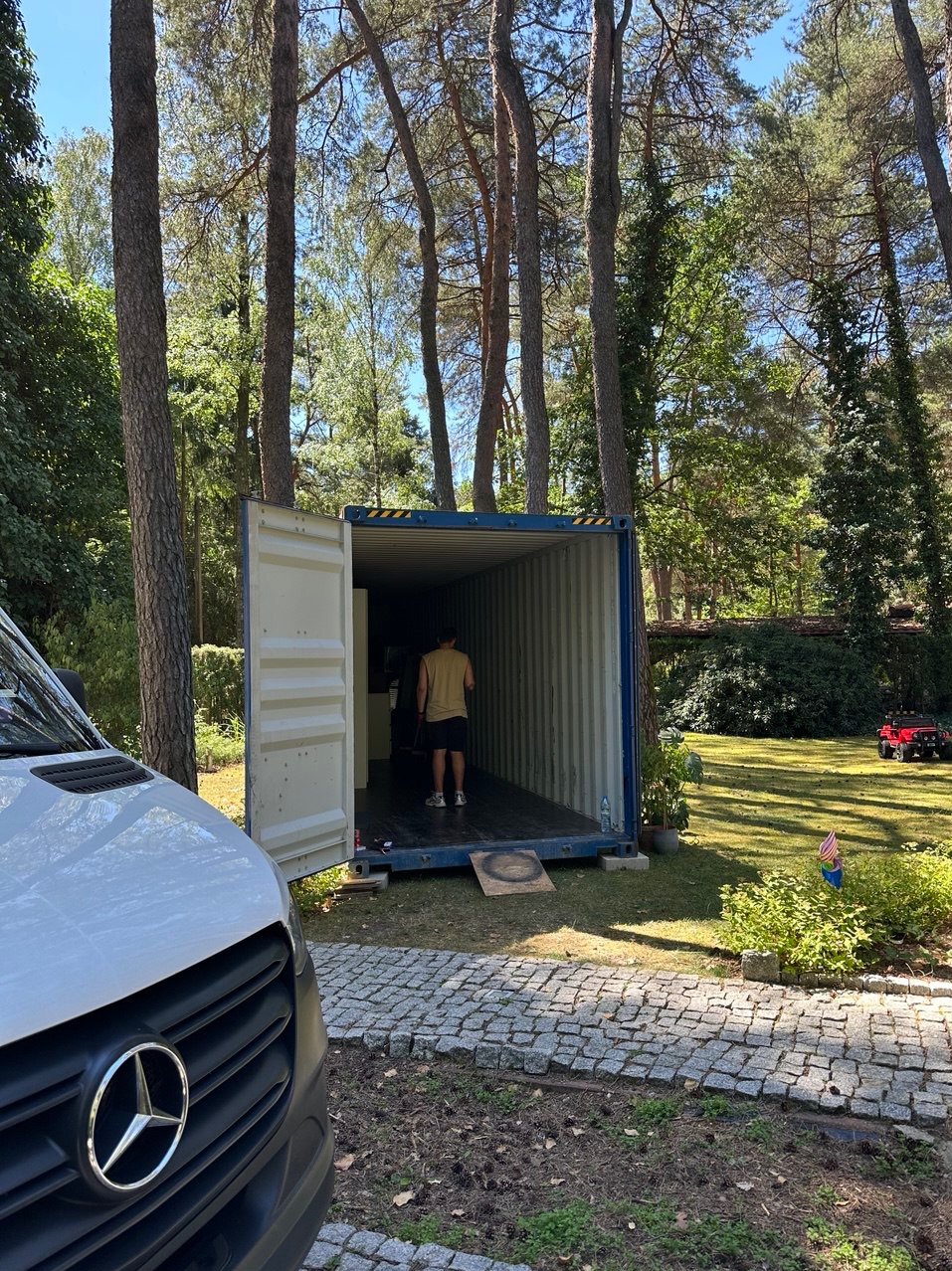 Otwarty kontener transportowy ustawiony na trawniku w otoczeniu drzew, osoba wchodzi do środka, obok zaparkowany biały bus marki Mercedes.