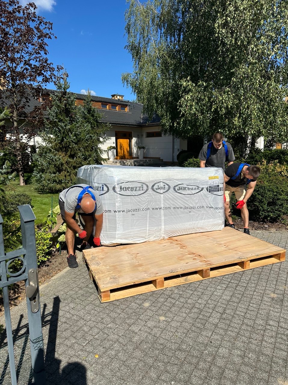 Trzech mężczyzn w czerwonych rękawicach przenosi zapakowane jacuzzi na drewnianej palecie przed domem z ogrodem w słoneczny dzień.