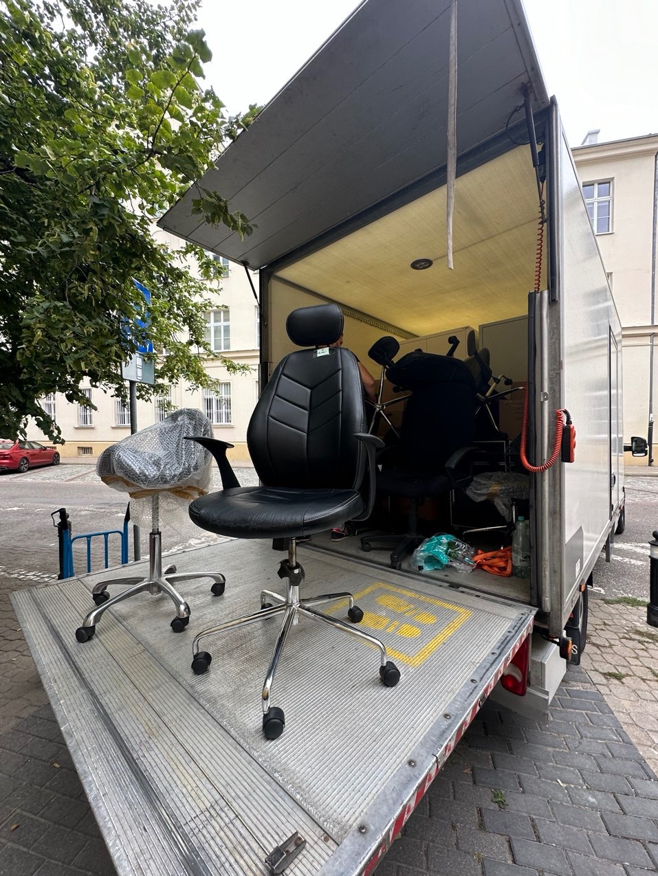 Czarne krzesło biurowe z zagłówkiem i krzesło obrotowe w folii na platformie załadunkowej busa z otwartymi drzwiami, widok z zewnątrz.