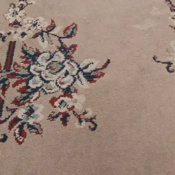Beżowy dywan z wzorem kwiatowym w odcieniach bieli, czerwieni i granatu, z widocznymi śladami użytkowania i niewielkimi plamami.