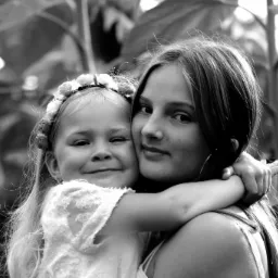 Czarno-białe zbliżenie uśmiechniętej dziewczynki w wianku obejmującej starszą nastolatkę na tle roślinności.