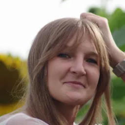 Portret młodej kobiety z blond włosami, uśmiechającej się, z ręką we włosach, na tle rozmytej zieleni i słonecznika.