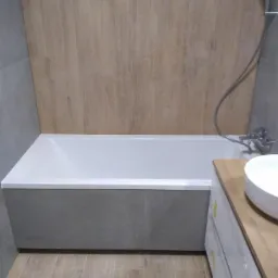 Nowoczesna łazienka z wanną obudowaną szarymi płytkami, drewnopodobną okładziną ścienną i umywalką nablatową na szafce z drewnianym blatem.