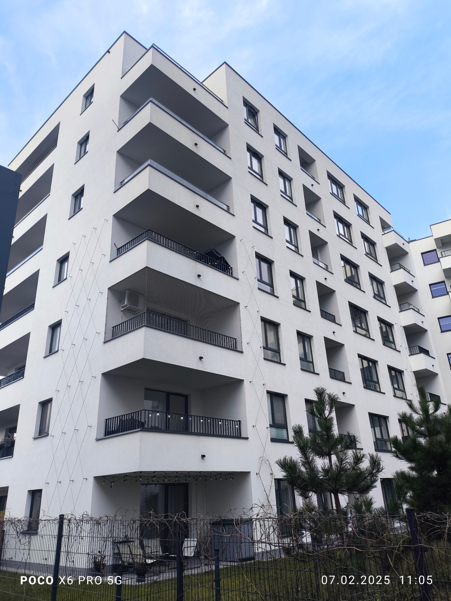 Nowoczesny, biały budynek wielorodzinny z balkonami, widoczny z dołu pod kątem. Na elewacji zainstalowano zabezpieczenia przeciw ptakom. U dołu drzewo iglaste.