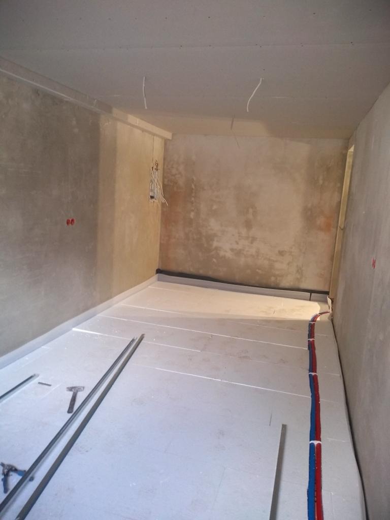 Wnętrze w trakcie prac: izolacja podłogi styropianem, widoczne rury instalacji grzewczej, surowe ściany i wystające przewody elektryczne.