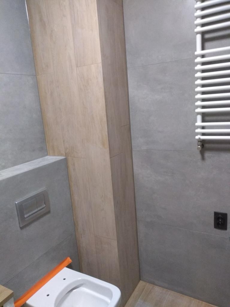 Wykończona łazienka z szarymi płytkami, drewnopodobną obudową stelaża WC, białym grzejnikiem drabinkowym i podtynkowym przyciskiem spłukującym.