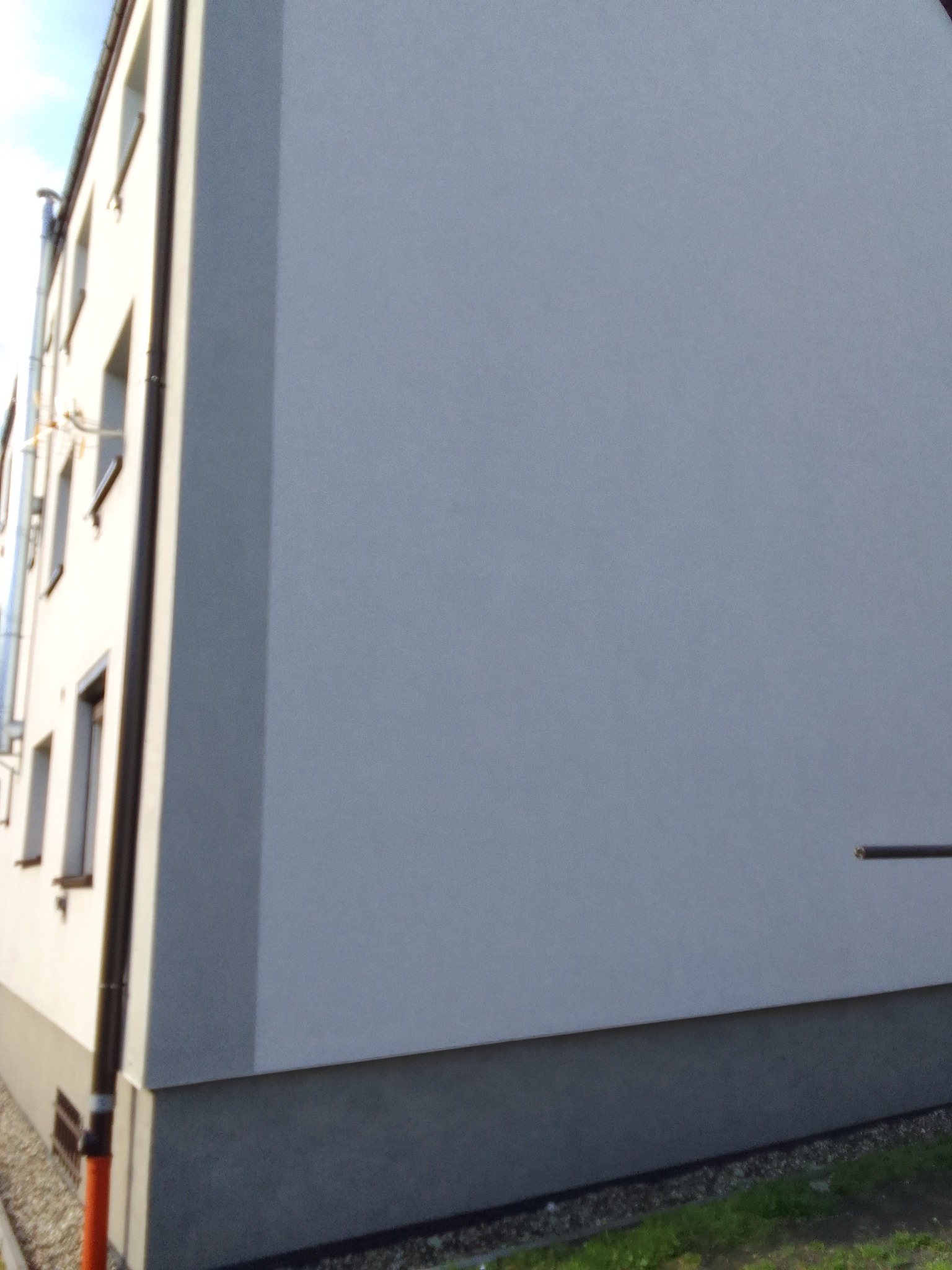 Budynek po ociepleniu z widoczną strukturą tynku zewnętrznego, brązowe rynny i okna, szara podmurówka i fragment trawnika.