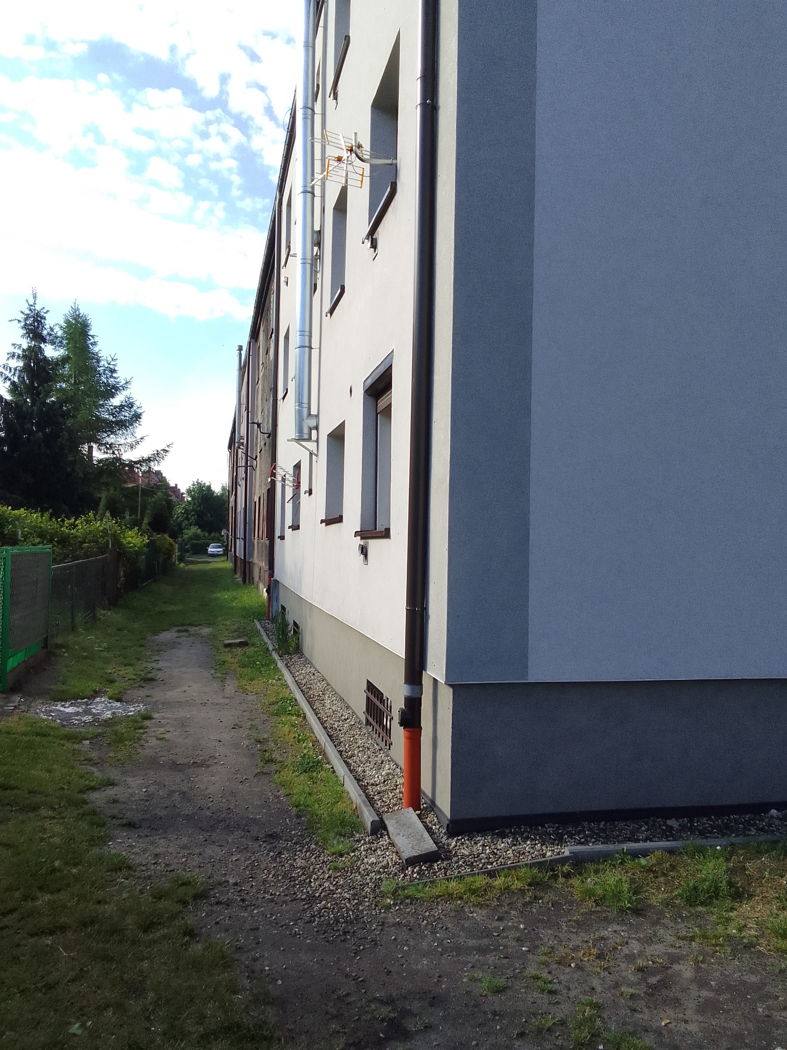 Boczny widok bloku mieszkalnego po renowacji elewacji, z widocznym nowym orynnowaniem i anteną telewizyjną na dachu, na tle zieleni i ścieżki.