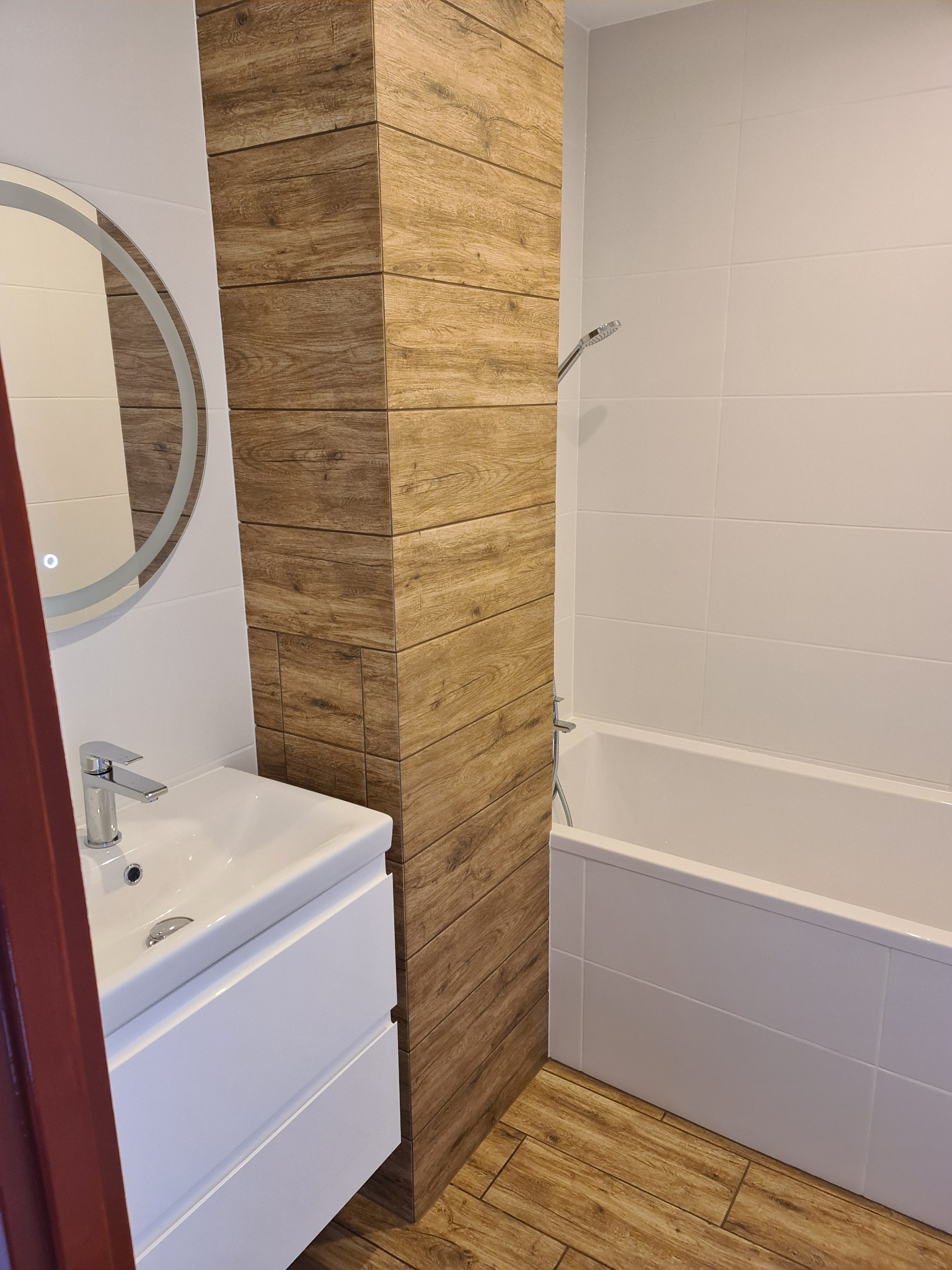 Nowoczesna łazienka z białą armaturą, okrągłym lustrem z podświetleniem LED i płytkami imitującymi drewno na podłodze i ścianie prysznicowej.