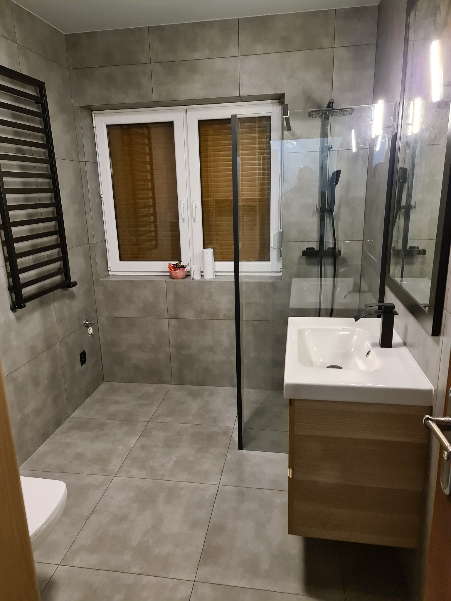 Wykończona łazienka w szarościach z drewnianą szafką pod umywalką, czarną armaturą i grzejnikiem drabinkowym, widoczna kabina prysznicowa ze szklaną ścianką.