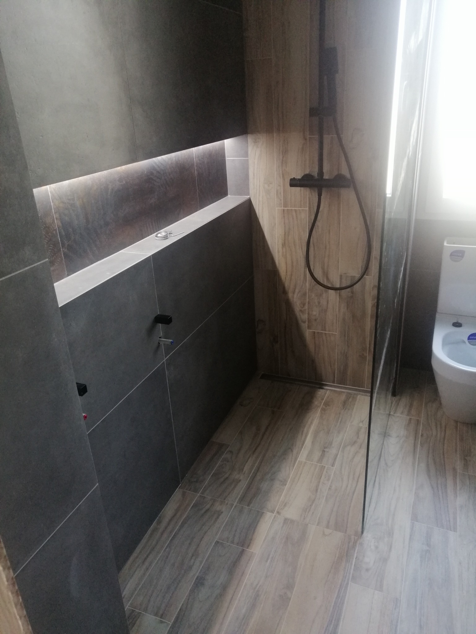 Nowoczesna łazienka z szarą zabudową, podświetlaną wnęką, drewnopodobnymi płytkami podłogowymi i czarną armaturą prysznicową za szklaną ścianką.