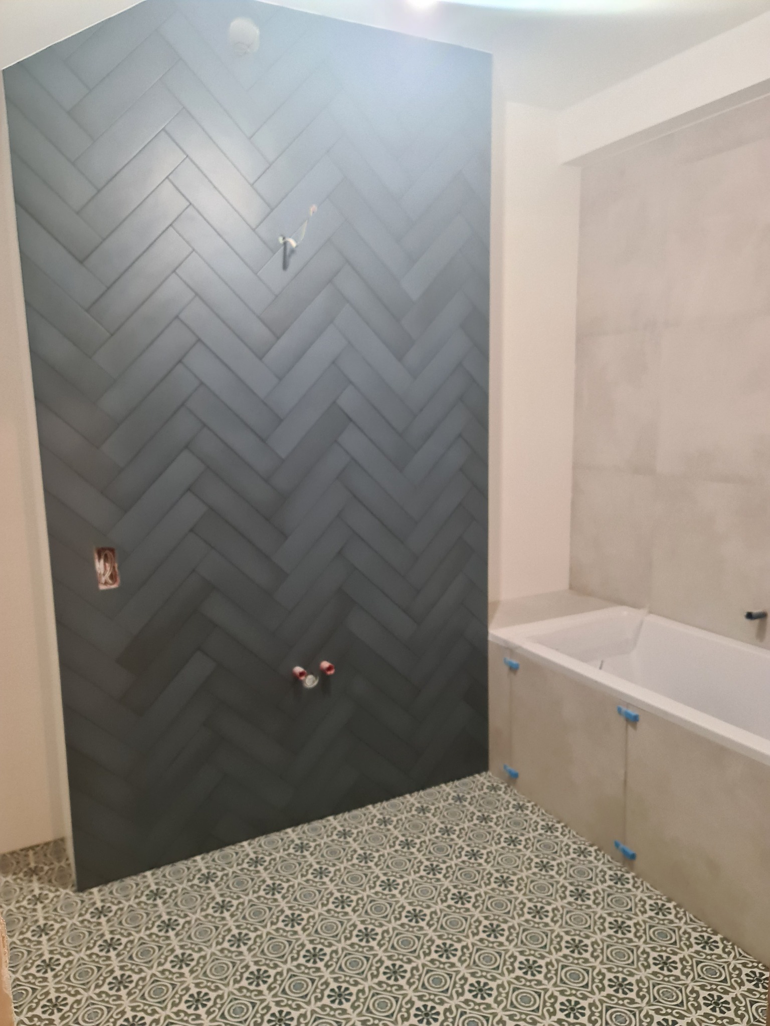 Ściana łazienki wyłożona ciemnymi, matowymi płytkami ułożonymi w jodełkę, widoczne wystające kable ze ściany i rury, na podłodze wzorzyste płytki, obok wanna w zabudowie.