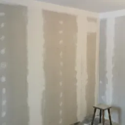 Pokój w trakcie szpachlowania, widoczne ściany z nałożoną gładzią w pasach, narzędzia pracy: wałek malarski, szpachelka i pojemnik z masą szpachlową, oraz drewniany stołek.