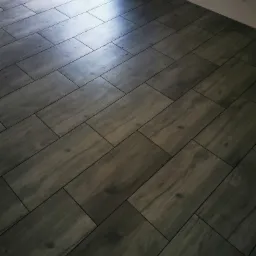 Ułożona pod kątem, ciemnoszara podłoga z płytek imitujących drewno, z widocznymi fugami i odbiciem światła w górnym rogu.