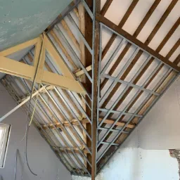 Poddasze w trakcie adaptacji: widoczne drewniane krokwie, metalowa konstrukcja nośna, fragmenty płyt kartonowo-gipsowych, okno i instalacja elektryczna.