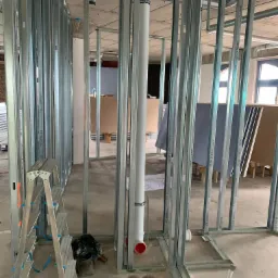 Stelaż z profili stalowych przygotowany pod montaż płyt kartonowo-gipsowych, widoczne instalacje sanitarne, drabina aluminiowa i narzędzia.