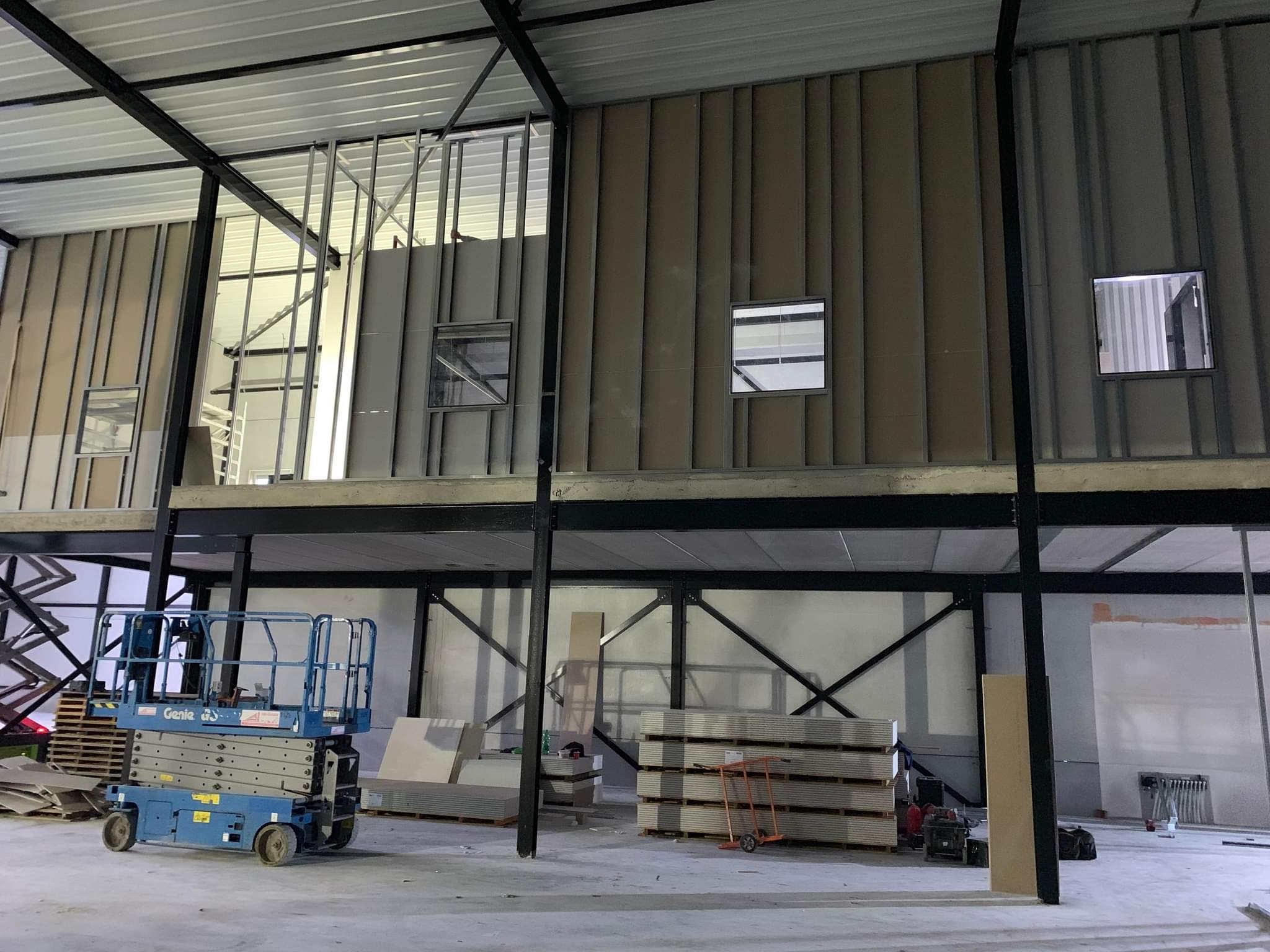 Wnętrze budynku w trakcie prac: montaż płyt warstwowych na ścianach z widocznymi oknami, konstrukcja stalowa, podnośnik nożycowy Genie i składowane płyty kartonowo-gipsowe.