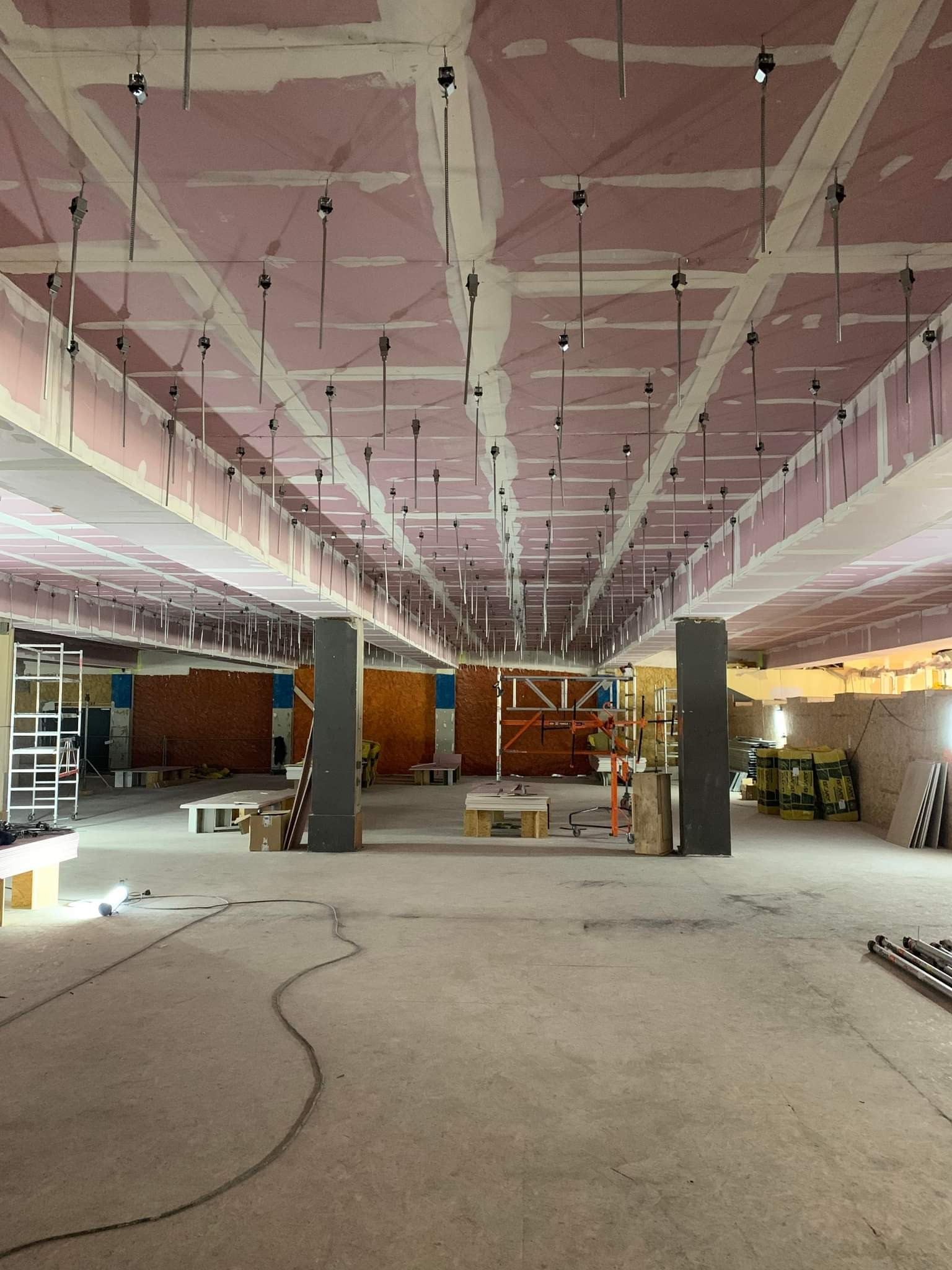 Rozległe wnętrze w trakcie montażu sufitu podwieszanego z płyt kartonowo-gipsowych, widoczne profile, zawiesia, rusztowania i materiały budowlane na podłodze, sufit w kolorze różowym.