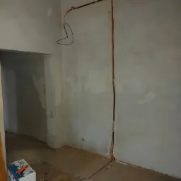 Wnętrze w trakcie remontu: bruzdy w ścianie i podłodze przygotowane pod instalację elektryczną, kabel zwisający ze ściany, karton z narzędziami obok wejścia do drugiego pomieszczenia.