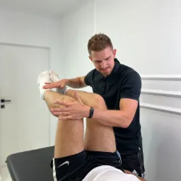 Mężczyzna w czarnej koszulce delikatnie rozciąga nogi pacjenta leżącego na czarnym stole do masażu w jasnym gabinecie.