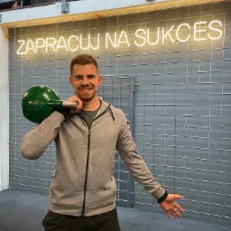 Uśmiechnięty mężczyzna w sportowym stroju trzyma zielony kettlebell na ramieniu, na tle siatki z neonowym napisem 'ZAPRACUJ NA SUKCES'.