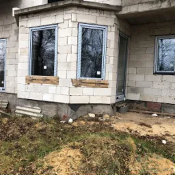 Surowy stan budynku z trzema oknami z folią ochronną i drzwiami wejściowymi, widoczne drewniane kliny pod oknami, teren wokół w trakcie prac ziemnych.