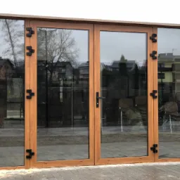 Zestaw dużych, przeszklonych drzwi dwuskrzydłowych z drewnopodobną ramą i czarnymi zawiasami, widok z zewnątrz na szarą kostkę brukową i odbijające się domy w szybach.